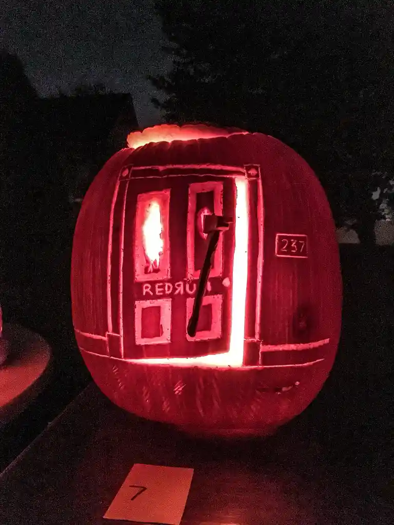 A carved pumpkin illuminated from within, depicting a door with an axe embedded in it, the word 'murder' carved below, and…