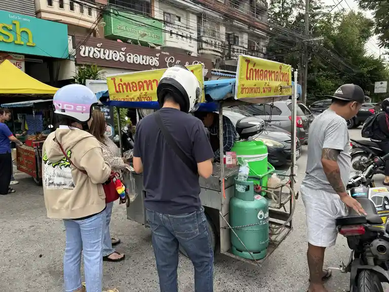 A street food vendor in Thailand serves customers from a cart equipped with a green propane tank, while people in helmets…