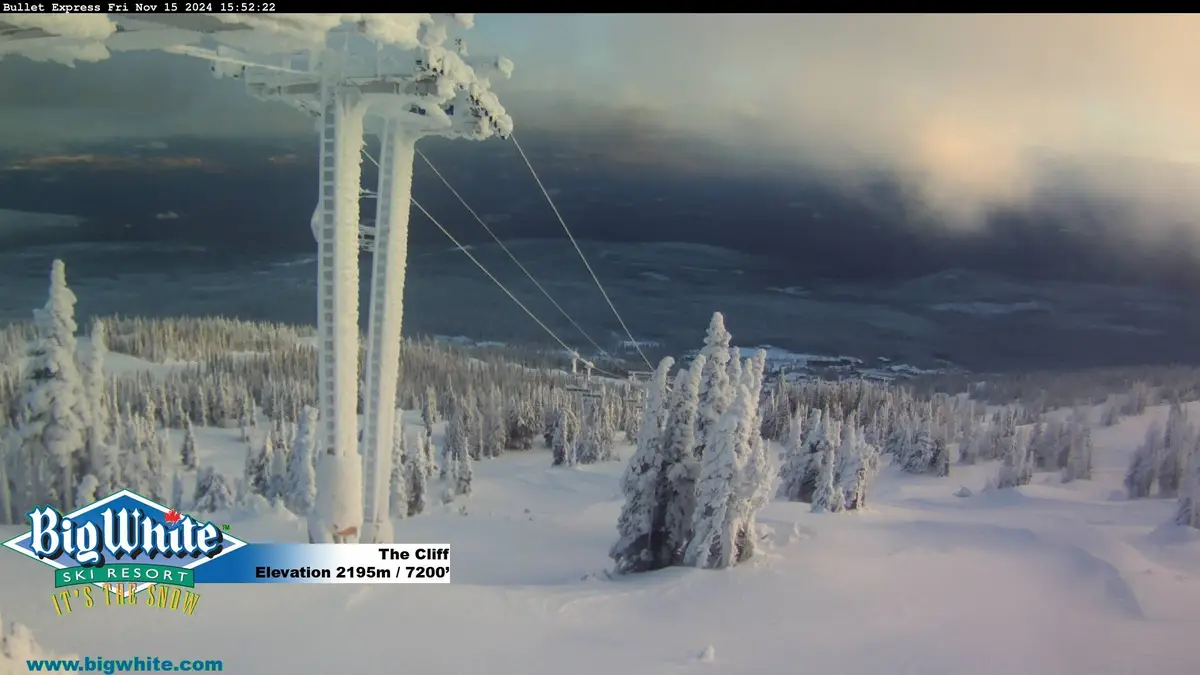 A snowy ski resort webcam view from Big White Ski Resort, showing a snow-covered chairlift tower and trees under a cloudy…