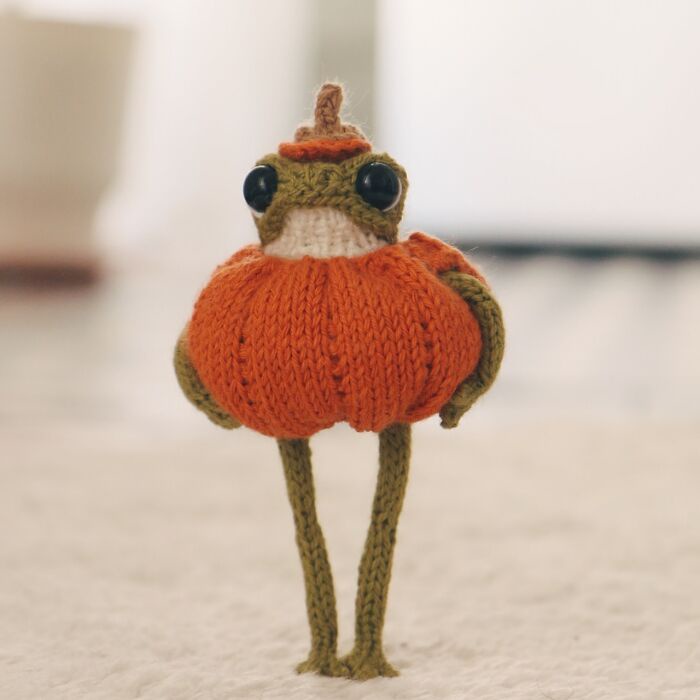 A small, hand-knitted frog figurine with large black eyes and green limbs is wearing an orange knitted pumpkin costume with…