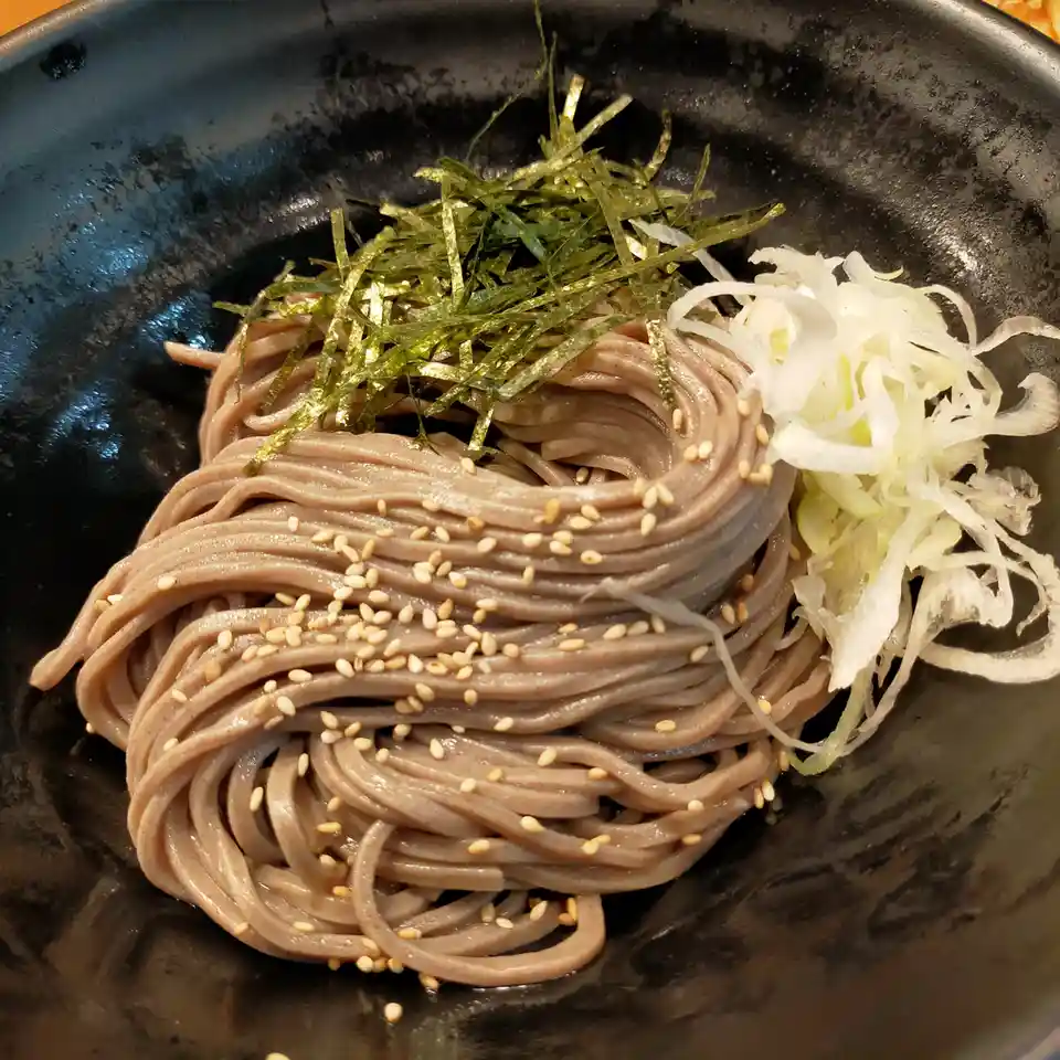A close-up photo of a dark ceramic bowl filled with brown soba noodles, topped with scattered white sesame seeds, a pile of…
