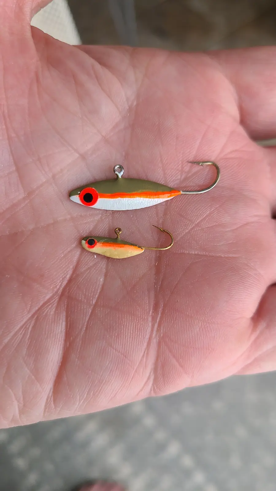 A person's open palm holds two small fishing lures, one larger and one smaller, both with red eyes and orange stripes. The…