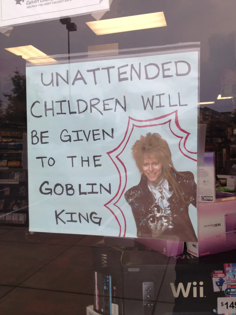 A hand-drawn sign taped to a store window reads 'UNATTENDED CHILDREN WILL BE GIVEN TO THE GOBLIN KING' with a red-bordered…