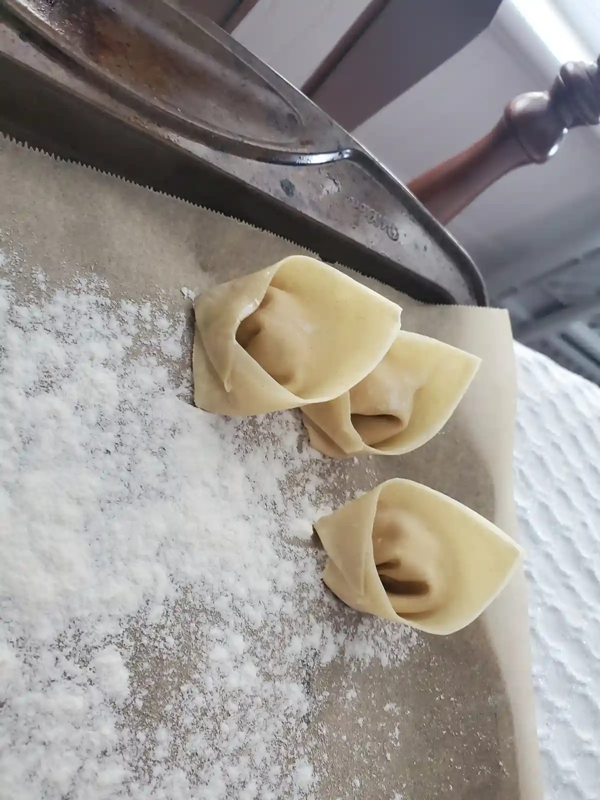 Three handmade dumplings sit on a floured metal baking sheet, their pleated edges resembling tear-streaked faces. The image…