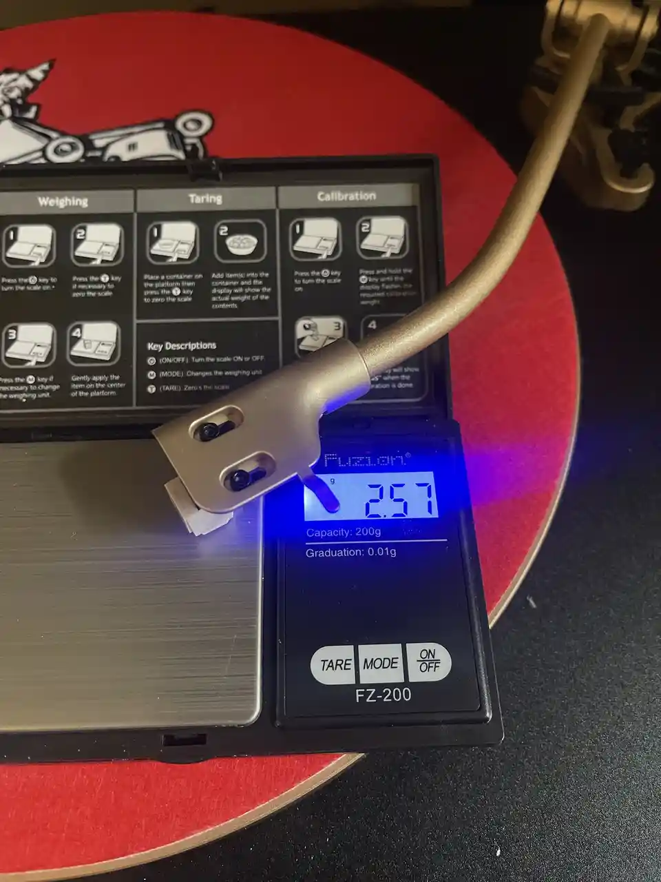 A digital kitchen scale displays '2.57' grams while a gold-colored turntable tonearm rests on its platform, which is placed…