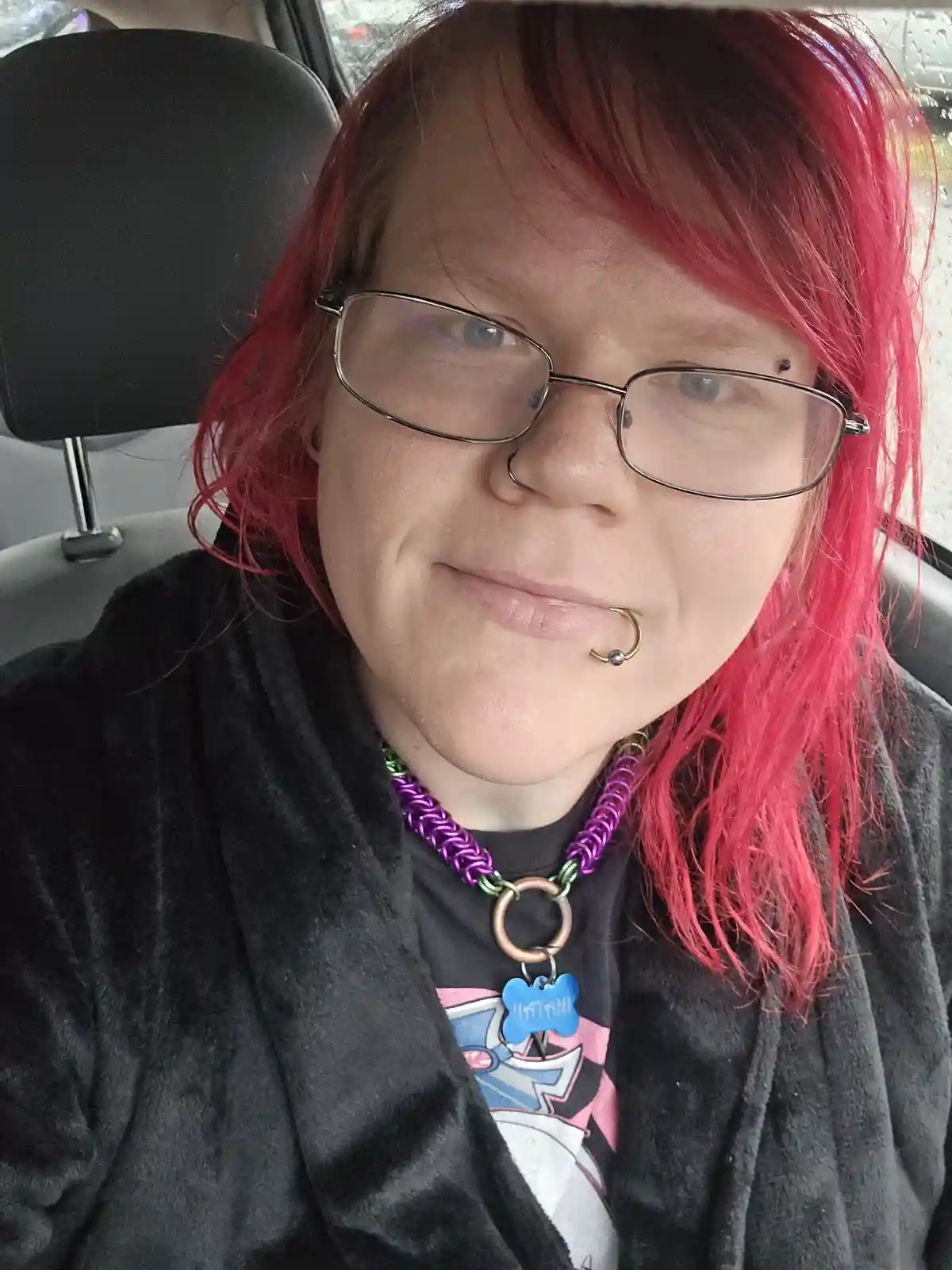 A person with bright pink hair, glasses, and a lip piercing sits in a car, wearing a black robe over a graphic t-shirt and a…