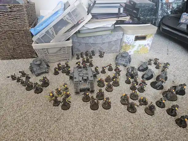 A large collection of painted miniature warhammer 40k space marine figures and vehicles are arranged on a carpeted floor…