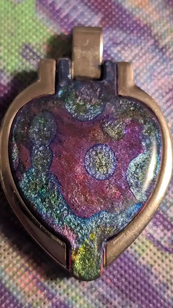 A close-up photo shows a handmade heart-shaped pendant with a bronze-colored metal frame and a colorful, iridescent glass or…