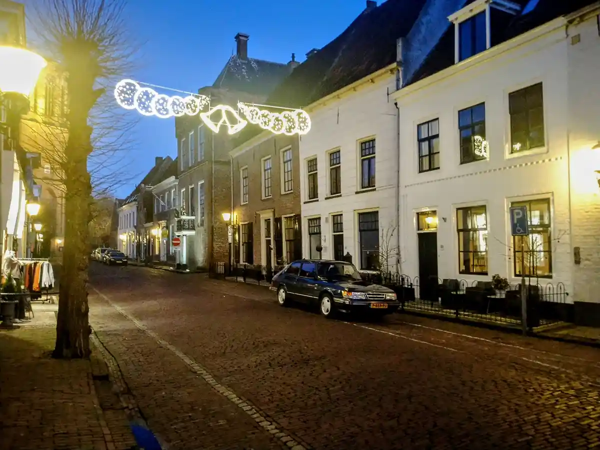 A cobblestone street at dusk with historic European-style buildings, a parked blue Saab car, and festive string lights…