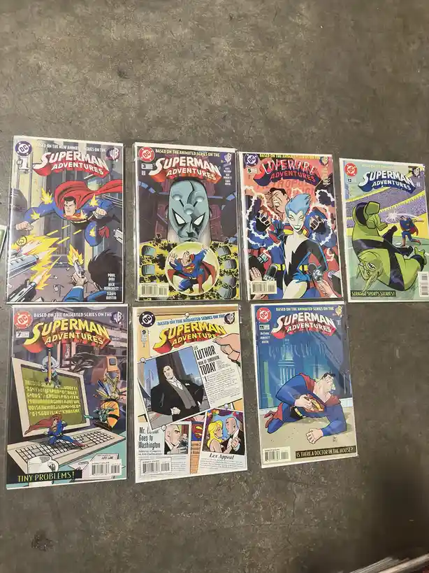 Seven Superman Adventures comic books are laid out on a gray concrete floor, each featuring different Superman storylines…