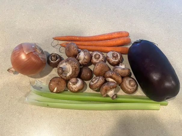 A flat lay of fresh vegetables including an onion, carrots, mushrooms, celery stalks, and an eggplant arranged on a light…