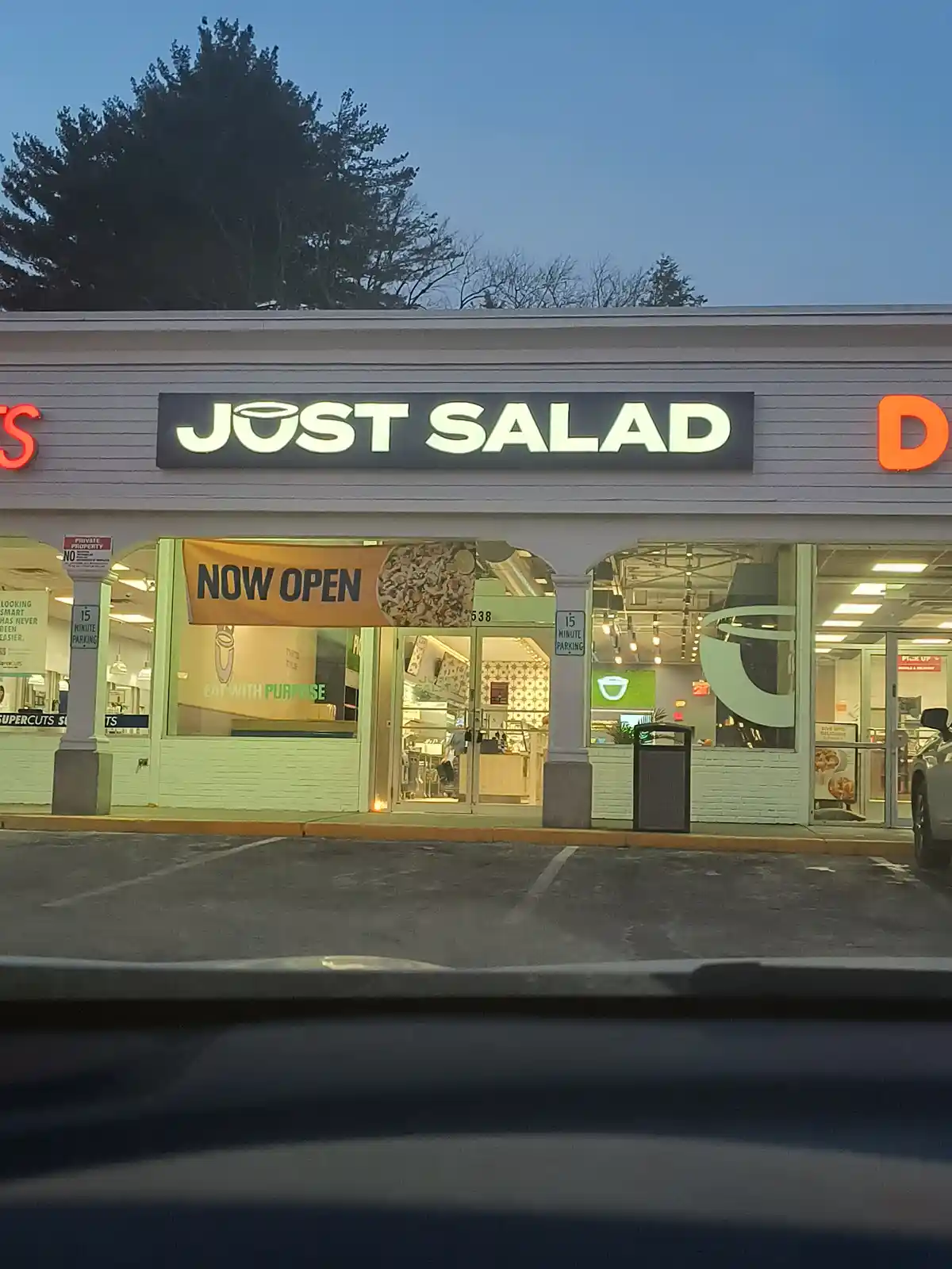 An exterior shot of a 'JUST SALAD' restaurant at dusk, viewed from inside a car. The store's sign features a salad bowl icon…