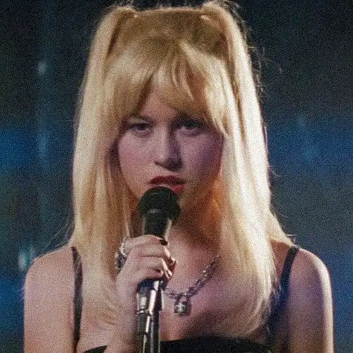 A woman with long blonde hair styled in two high pigtails holds a microphone, looking directly at the camera with a serious…