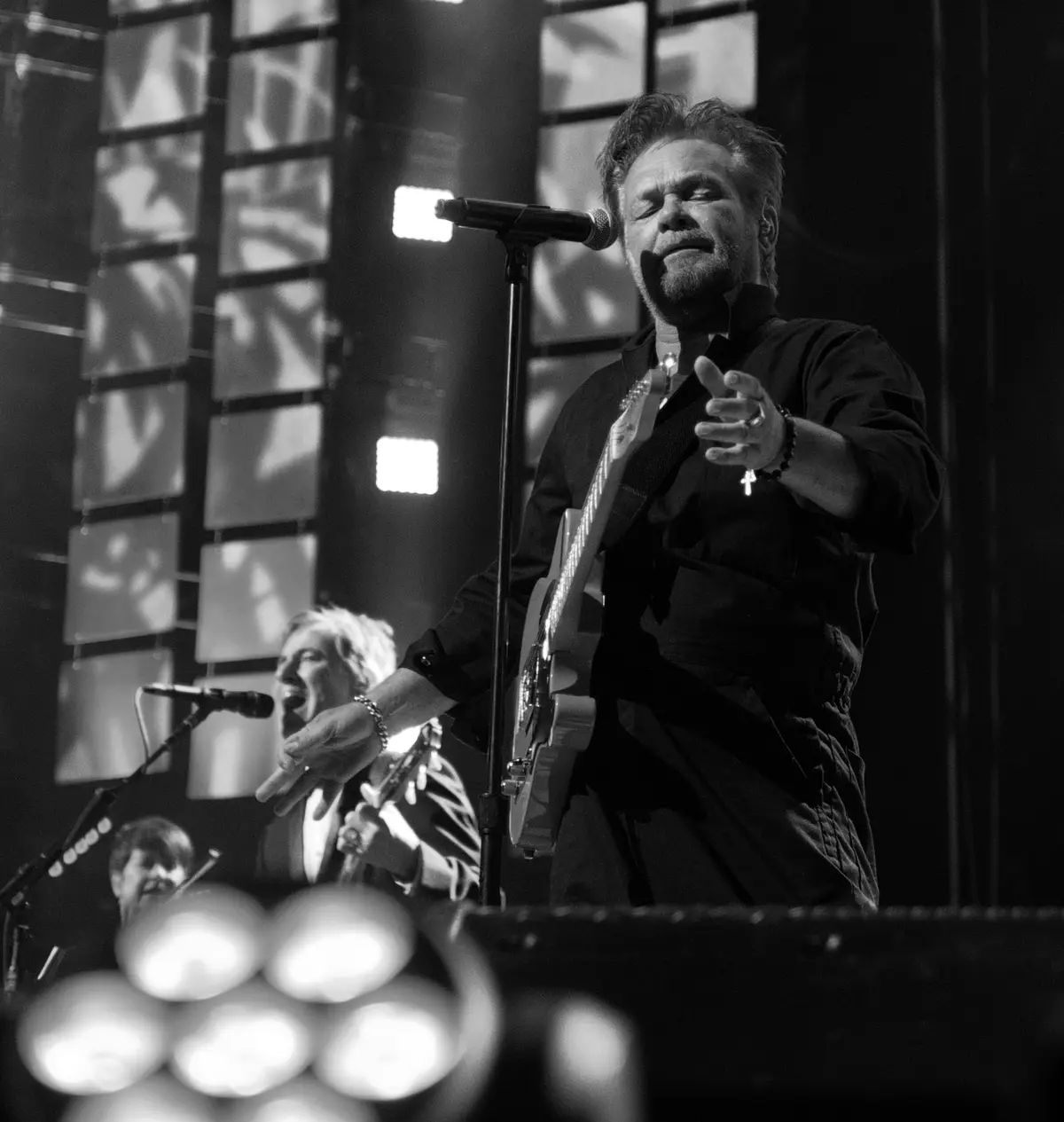 A black and white photo of a male musician with a beard, wearing a dark shirt and holding an electric guitar, singing into a…