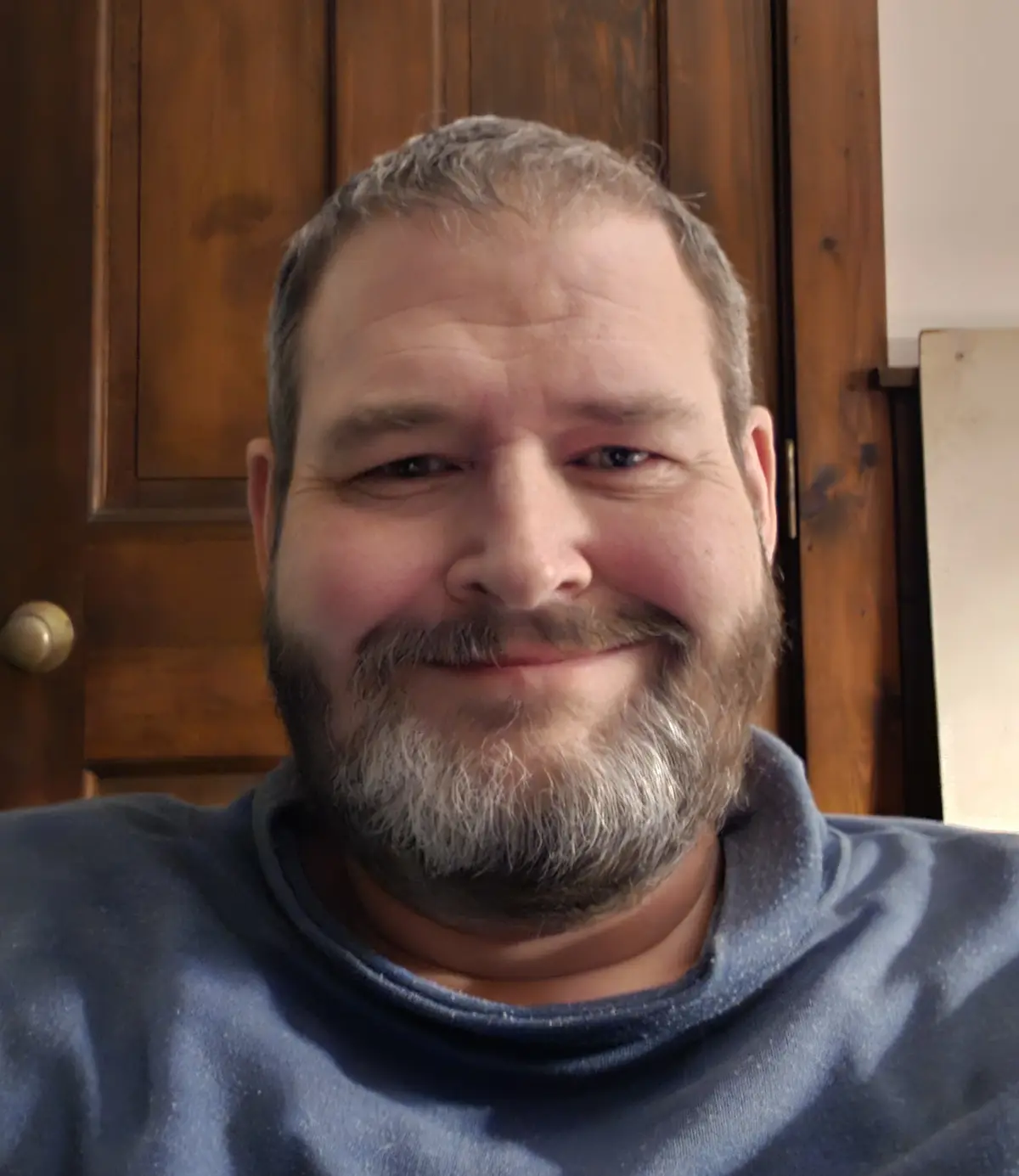 A close-up selfie of a middle-aged man with a graying beard and short hair, wearing a blue t-shirt, smiling slightly while…