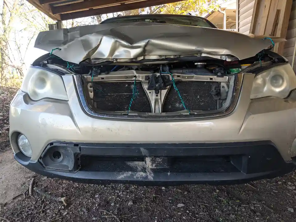 A silver car's front end is severely damaged, with the hood crumpled and the bumper missing, and the engine bay exposed…