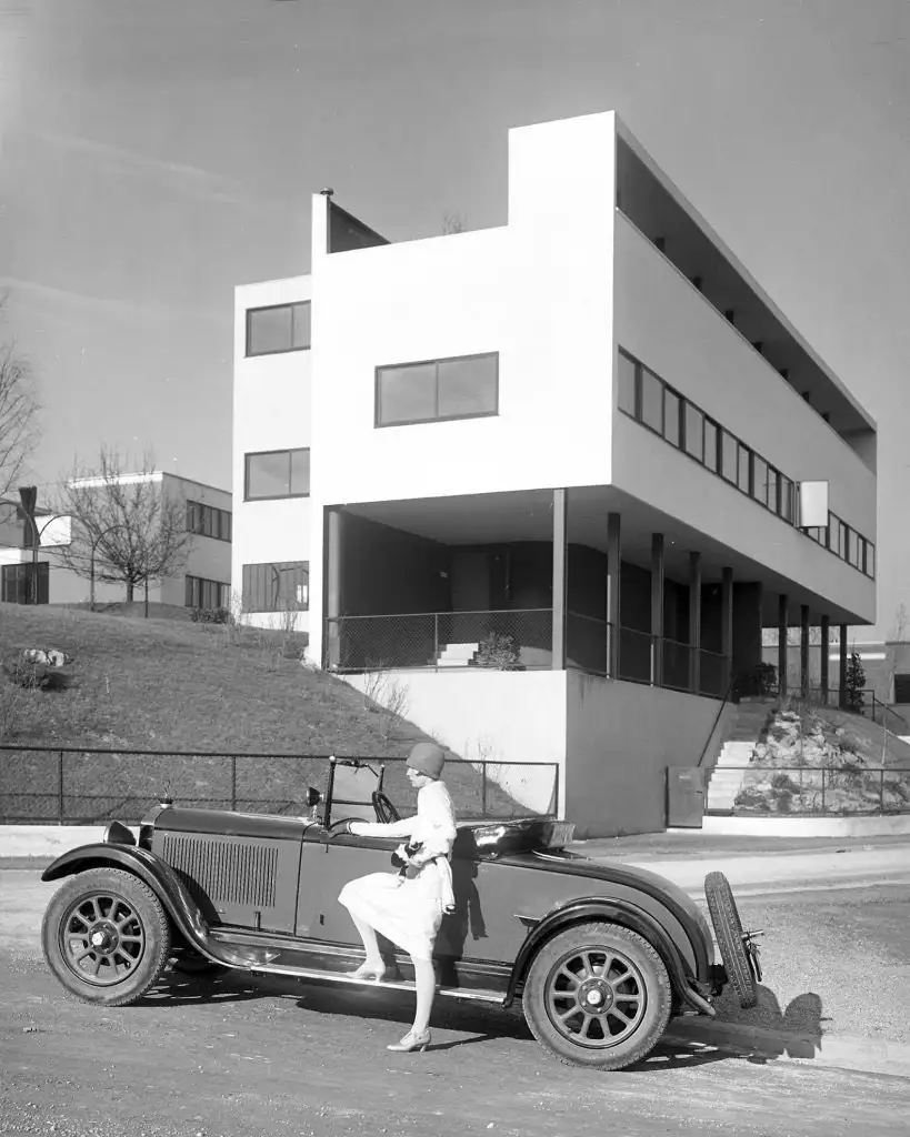A black-and-white photo shows a woman in a 1920s-style dress and cloche hat stepping out of a vintage convertible parked in…