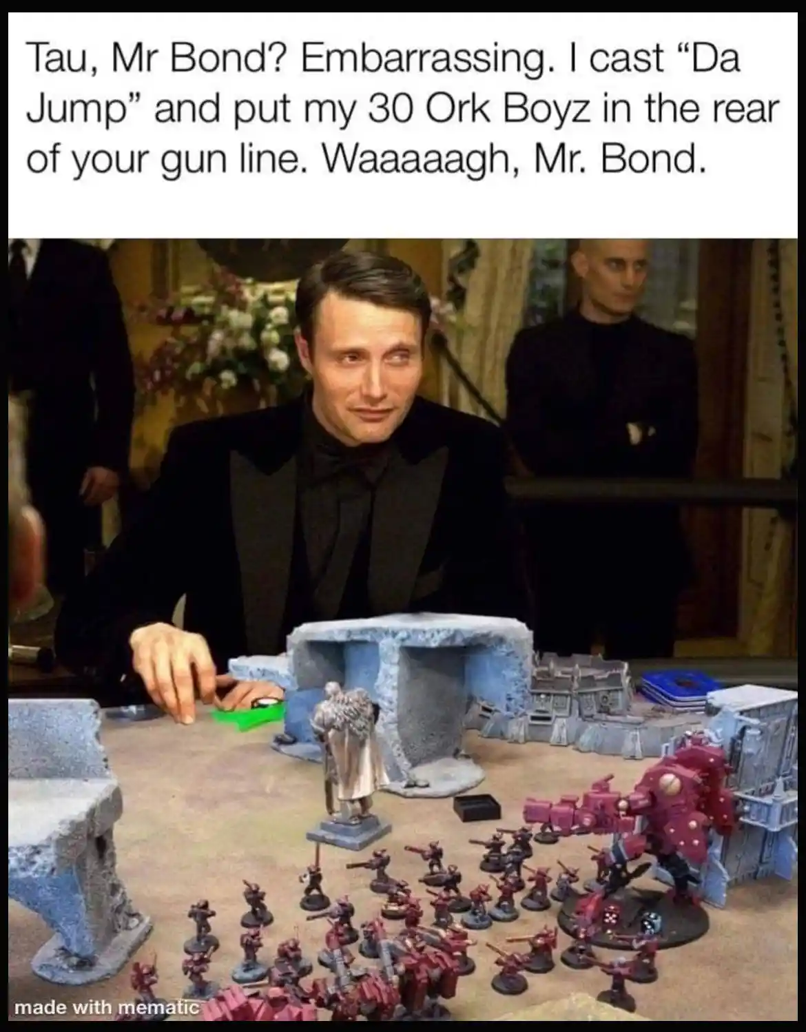 A man in a black tuxedo sits at a table with miniature wargaming figures, looking smugly sideways. The table has blue…