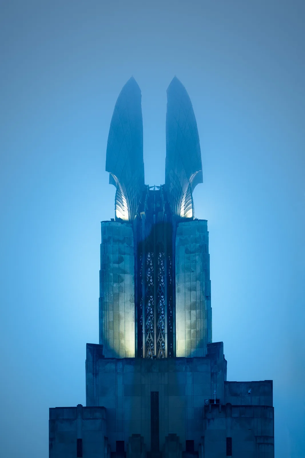 A tall, symmetrical building with two large, wing-like structures at its peak, illuminated against a blue sky. The overlaid…