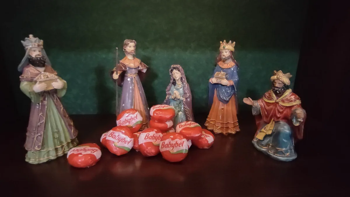 A nativity scene figurine set featuring Mary, Joseph, and the three wise men, with a pile of Babybel cheese wheels placed in…