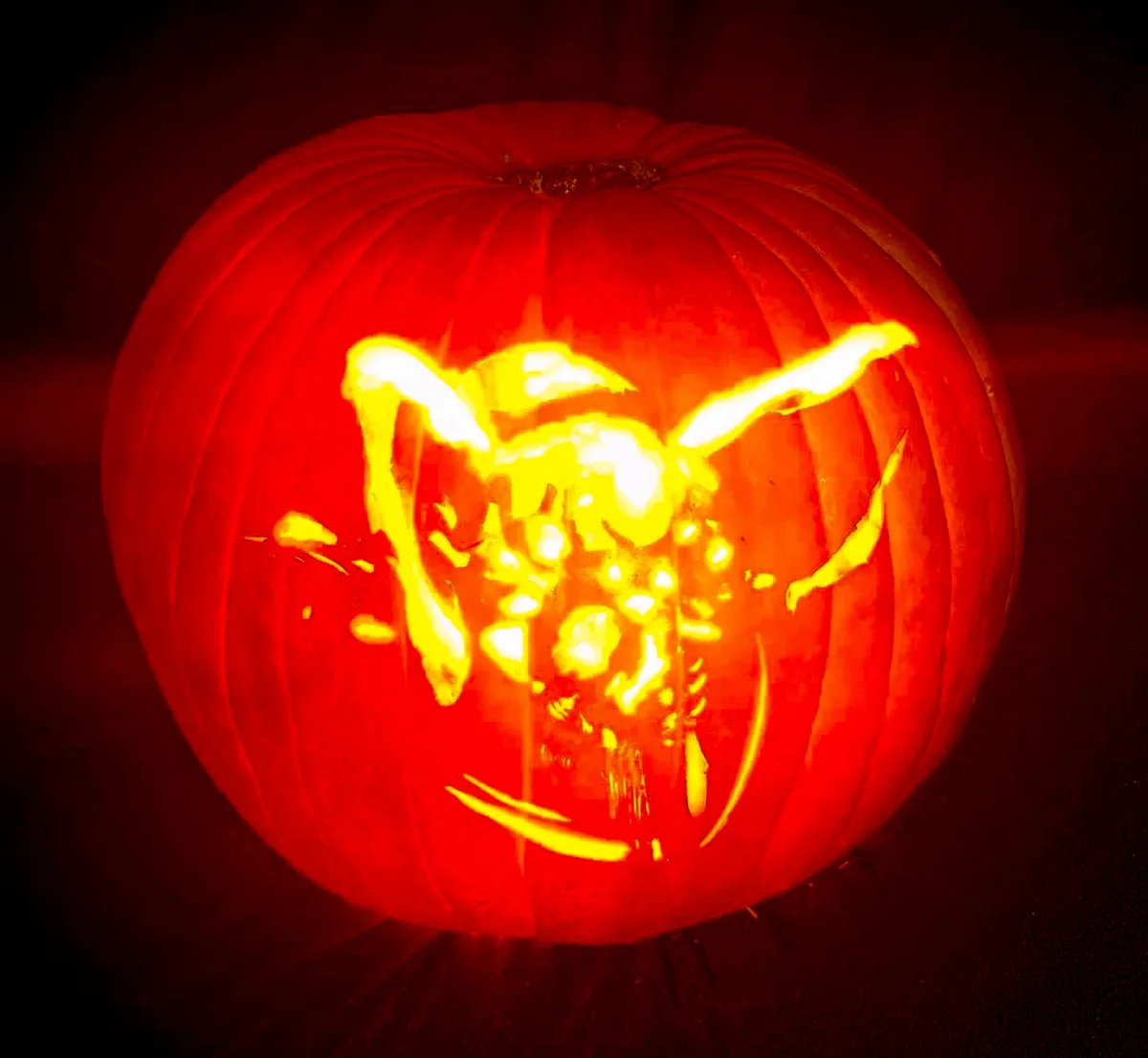 A carved jack-o'-lantern with a glowing, crudely cut face resembling a cartoonish, possibly demonic or monstrous figure with…