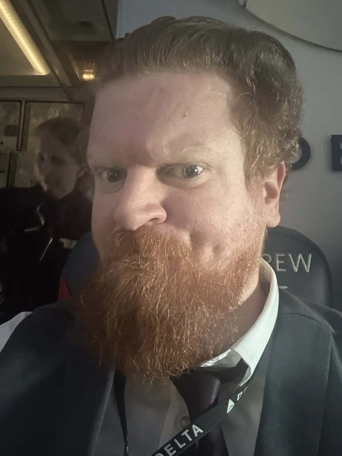A man with a red beard and crew uniform, including a Delta lanyard and 'CREW' chair, stares intensely at the camera with a…