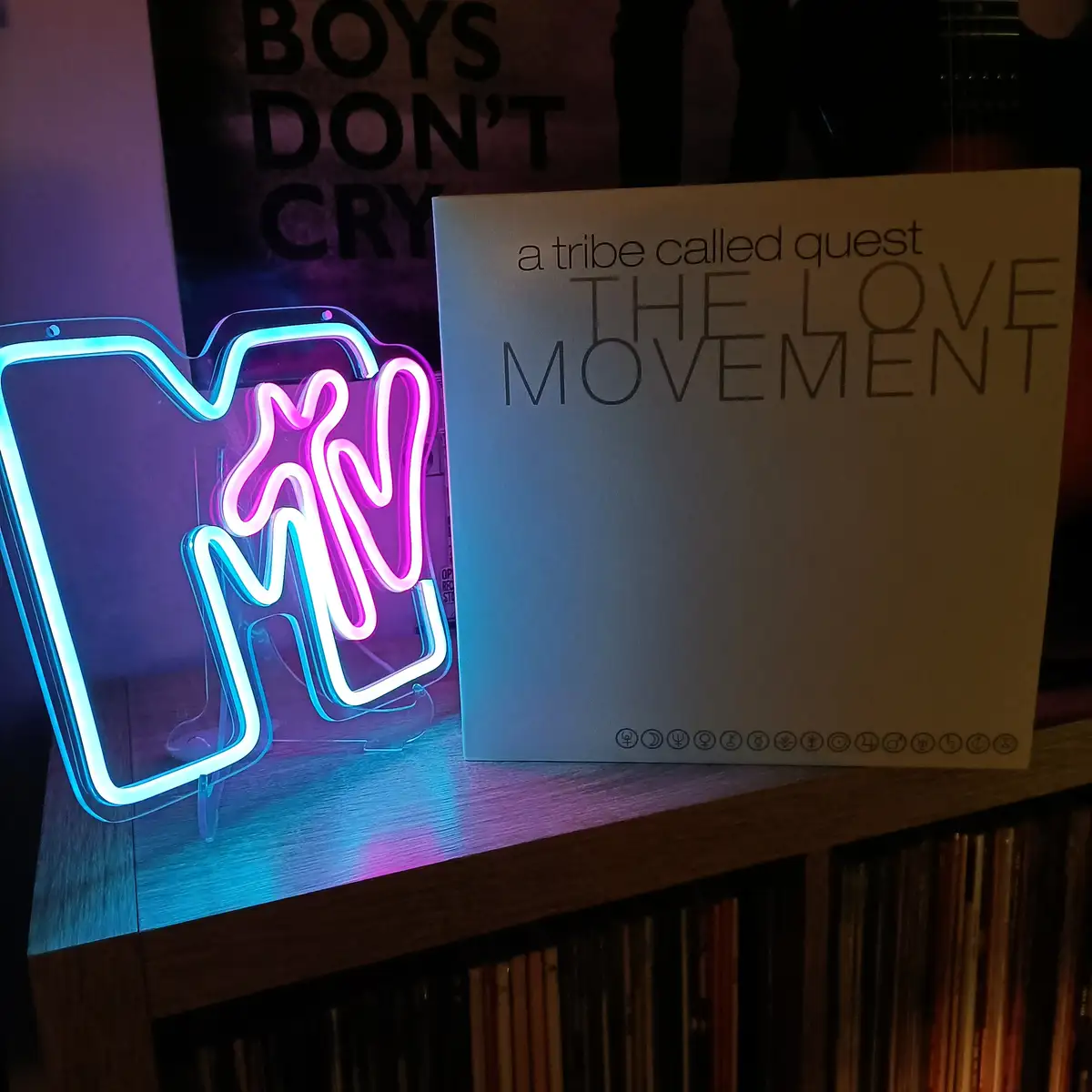 A neon sign shaped like a stylized 'M' with blue and pink lights sits next to a physical copy of the album 'The Love…