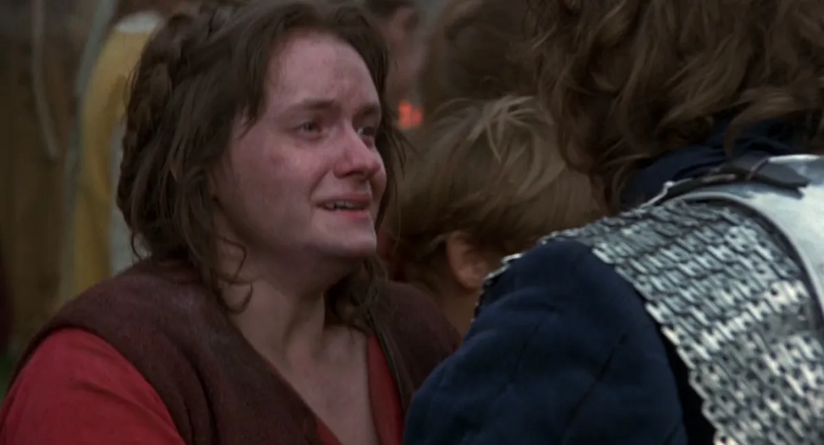 A woman in a red shirt and brown vest, visibly crying with tears on her face, looks up at a person in chainmail armor. The…