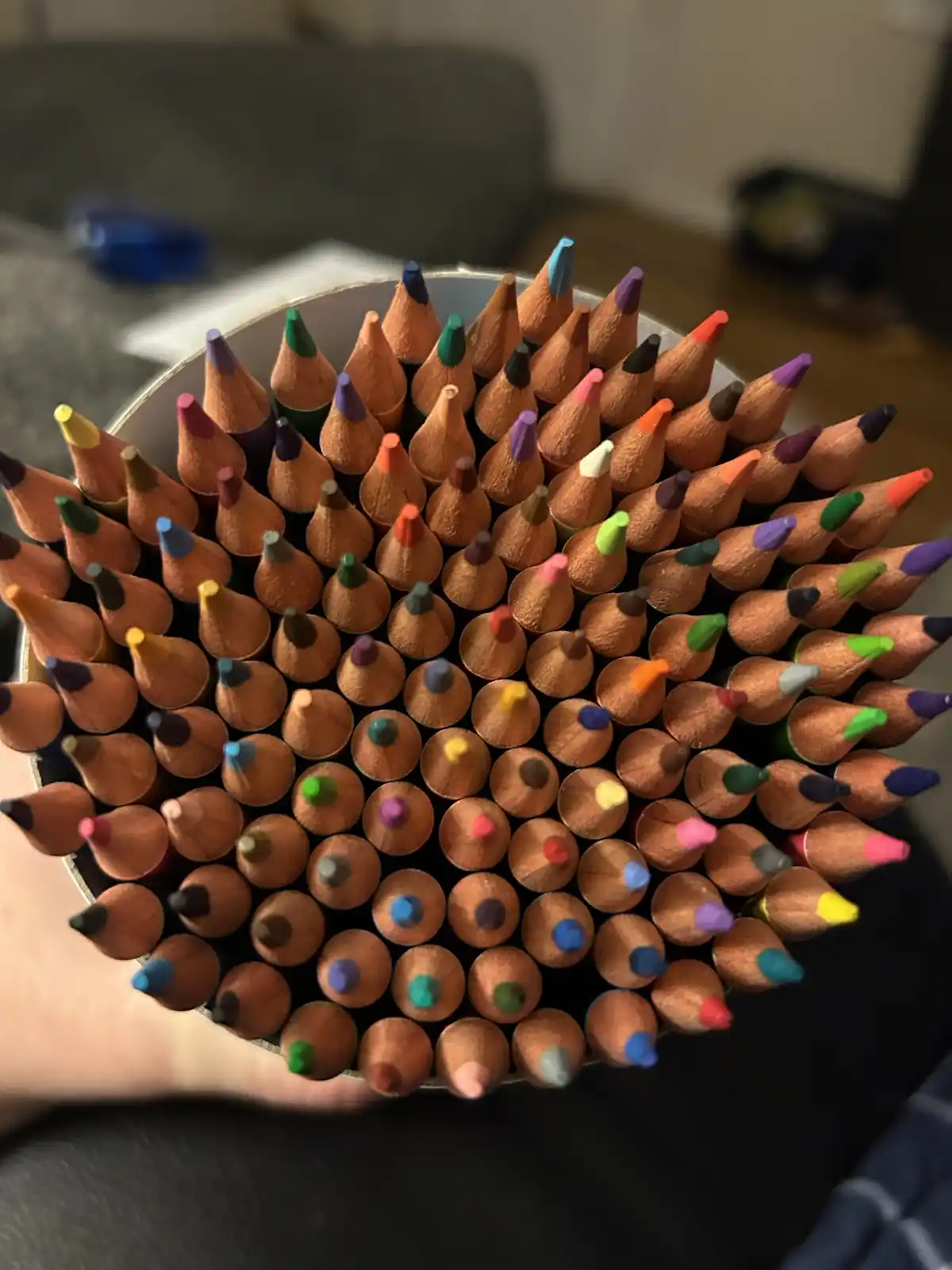 A top-down view shows a hand holding a cylindrical container overflowing with dozens of sharpened colored pencils, their…