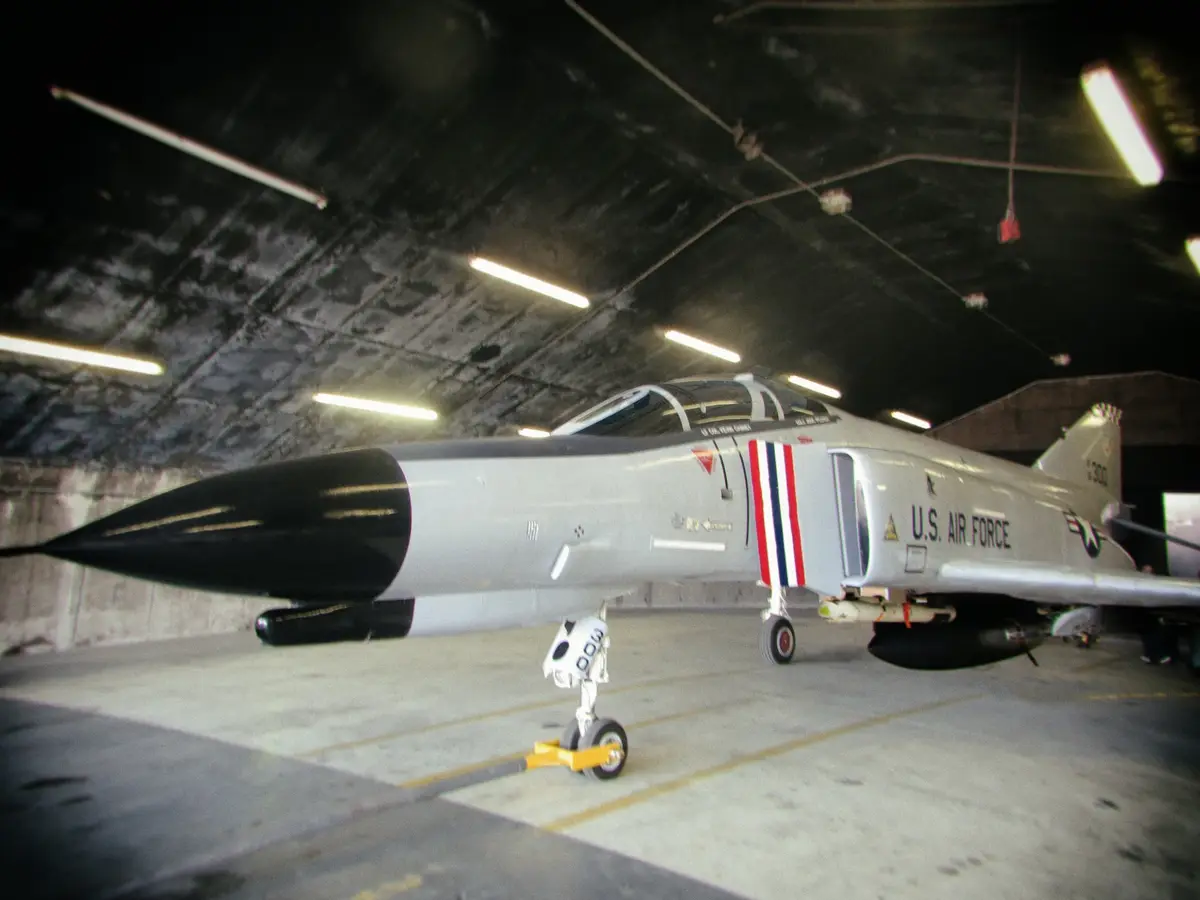 A U.S. Air Force fighter jet, likely a F-4 Phantom II, is parked inside a dimly lit concrete hangar with overhead…