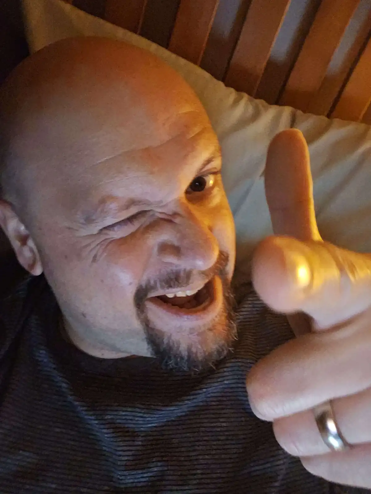 A bald man with a goatee lies in bed, winking one eye and giving a thumbs-up with his right hand, which has a ring on the…