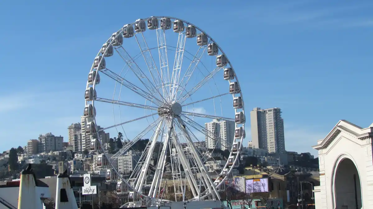 A large white Ferris wheel with enclosed gondolas stands against a clear blue sky, with a cityscape of buildings on hills in…