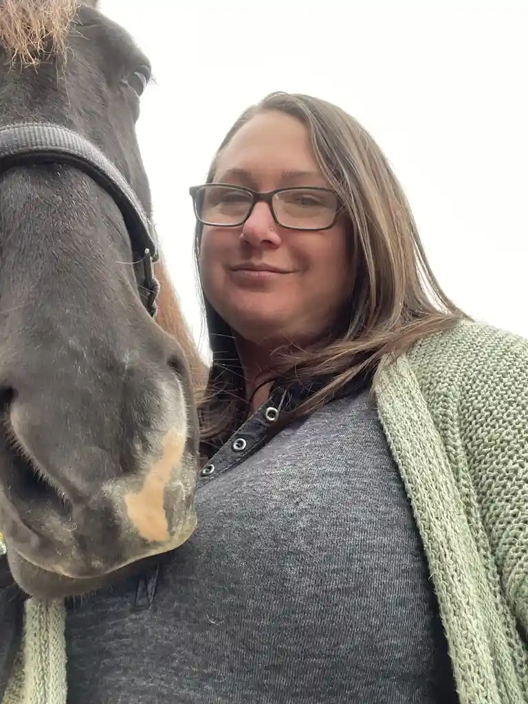 A woman with glasses and a green cardigan smiles while taking a selfie with a dark horse wearing a halter; the horse's…