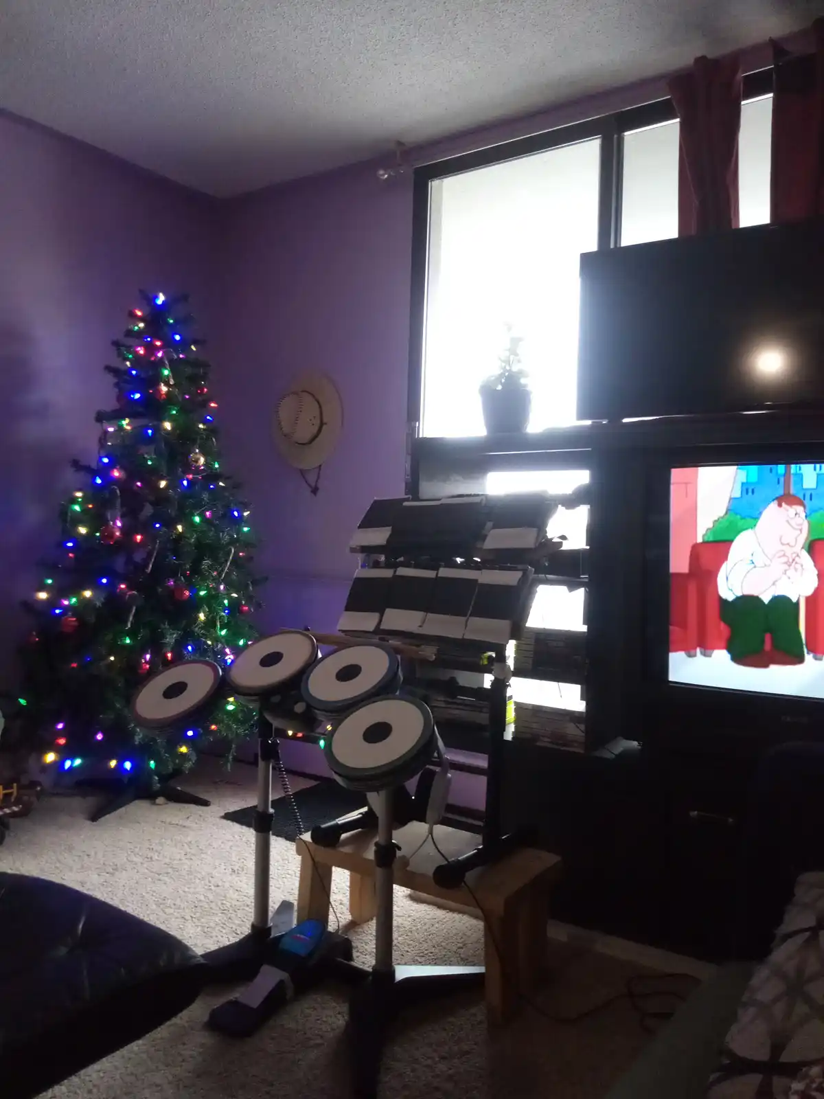 A room with a lit-up Christmas tree on the left and a drum set in the center, with a TV on the right showing the cartoon…