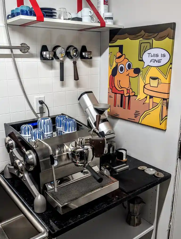 A coffee station with a professional espresso machine, grinder, and blue-and-white ceramic cups, next to a framed cartoon of…