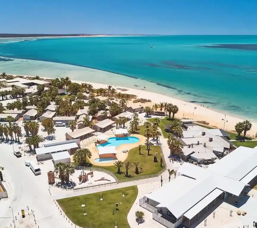 an aerial photo shows a resort with white-roofed buildings, palm trees, and a blue swimming pool near a sandy beach with…