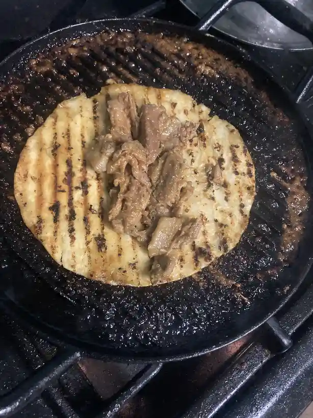 A close-up photo shows a round, grilled tortilla on a black cast-iron grill pan, with char marks visible. On top of the…
