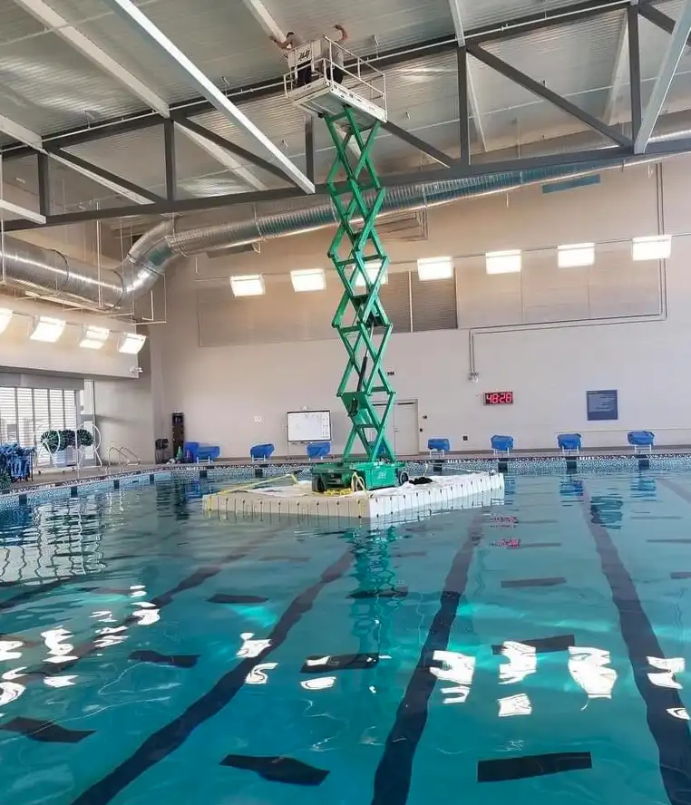 An indoor swimming pool with clear blue water and black lane lines. A green scissor lift is positioned in the middle of the…