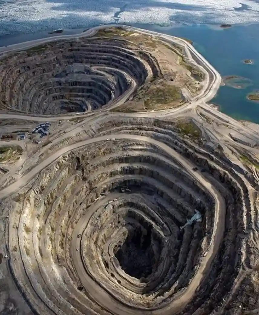 An aerial view shows two massive, terraced open-pit mines with concentric rings of earth and dirt roads winding around their…