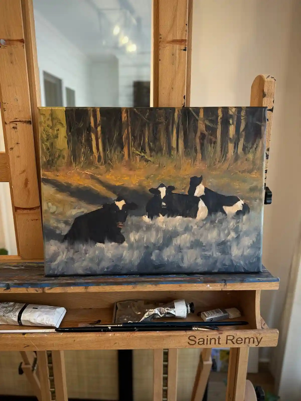 A painting of three black-and-white cows resting in a field under a forest, displayed on a wooden easel labeled 'Saint…