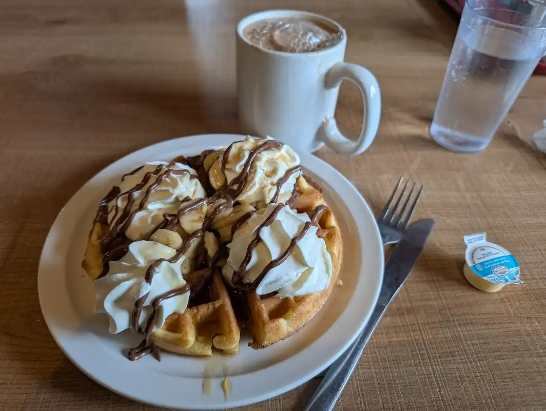 A plate of waffles topped with whipped cream, sliced bananas, and chocolate syrup sits on a wooden table next to a white mug…