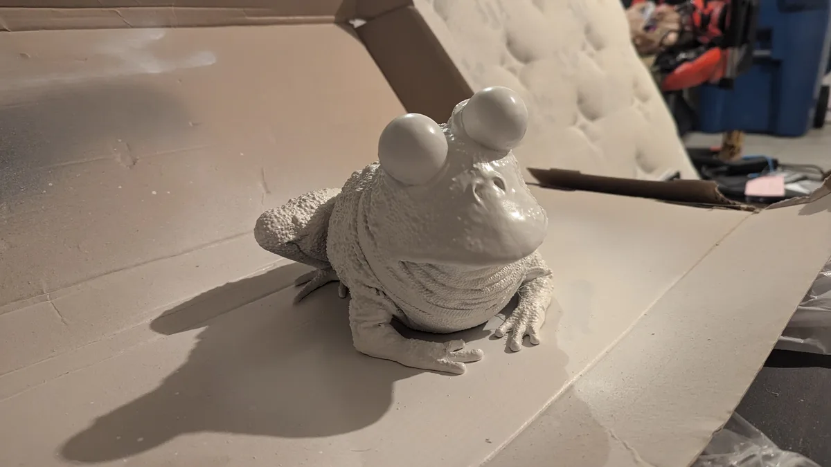 A white-painted frog statue with large, round eyes sits on a cardboard surface, looking directly at the camera. The overlaid…