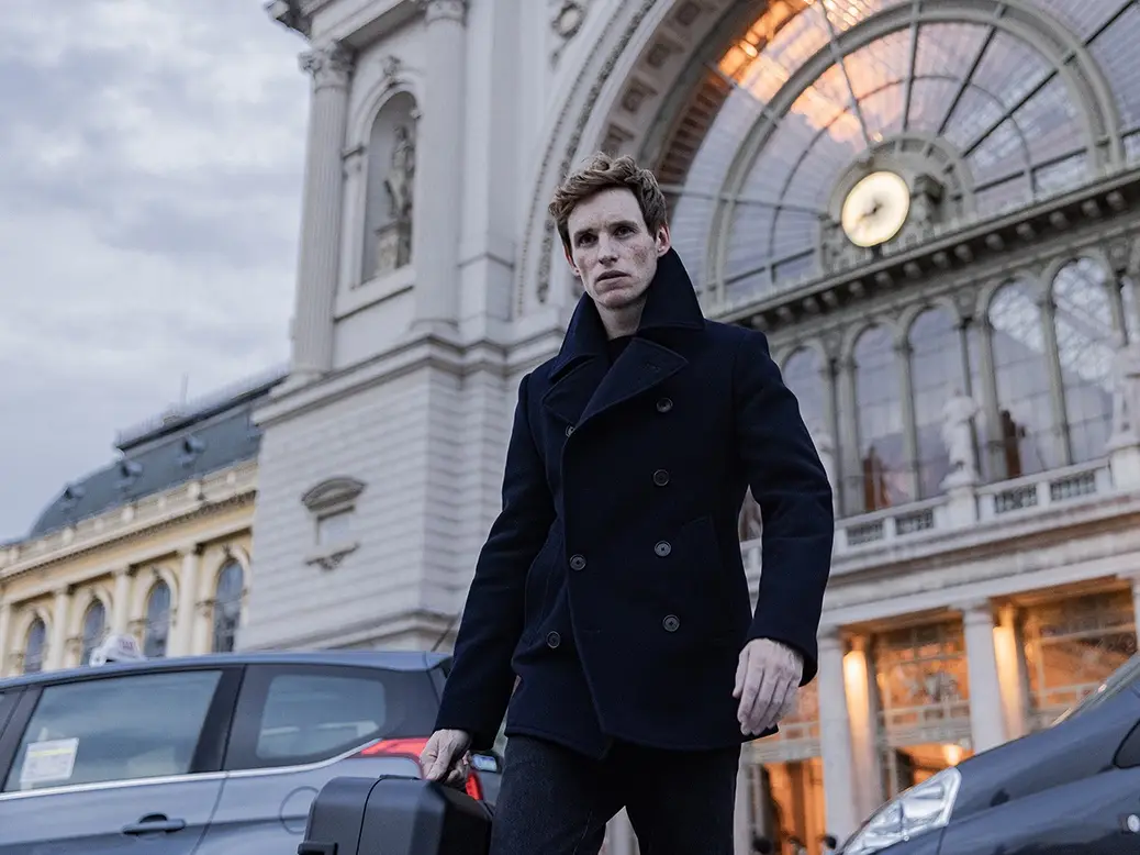 A man in a dark peacoat stands in front of a grand, ornate building with a large clock and glass dome, holding a suitcase…