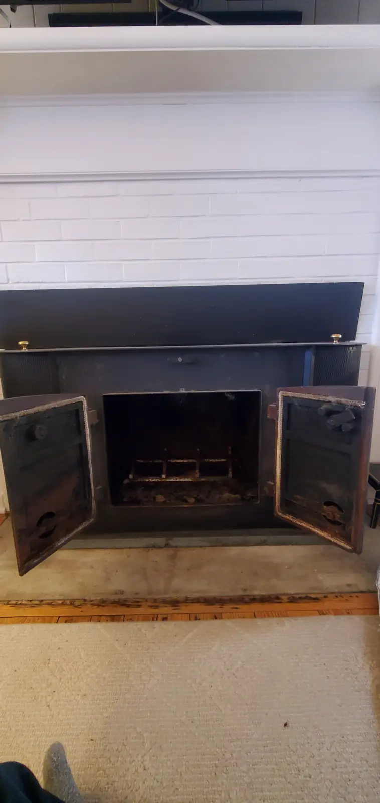 A photo shows an empty, open fireplace with its doors swung wide, revealing a dark, soot-stained interior with a metal…