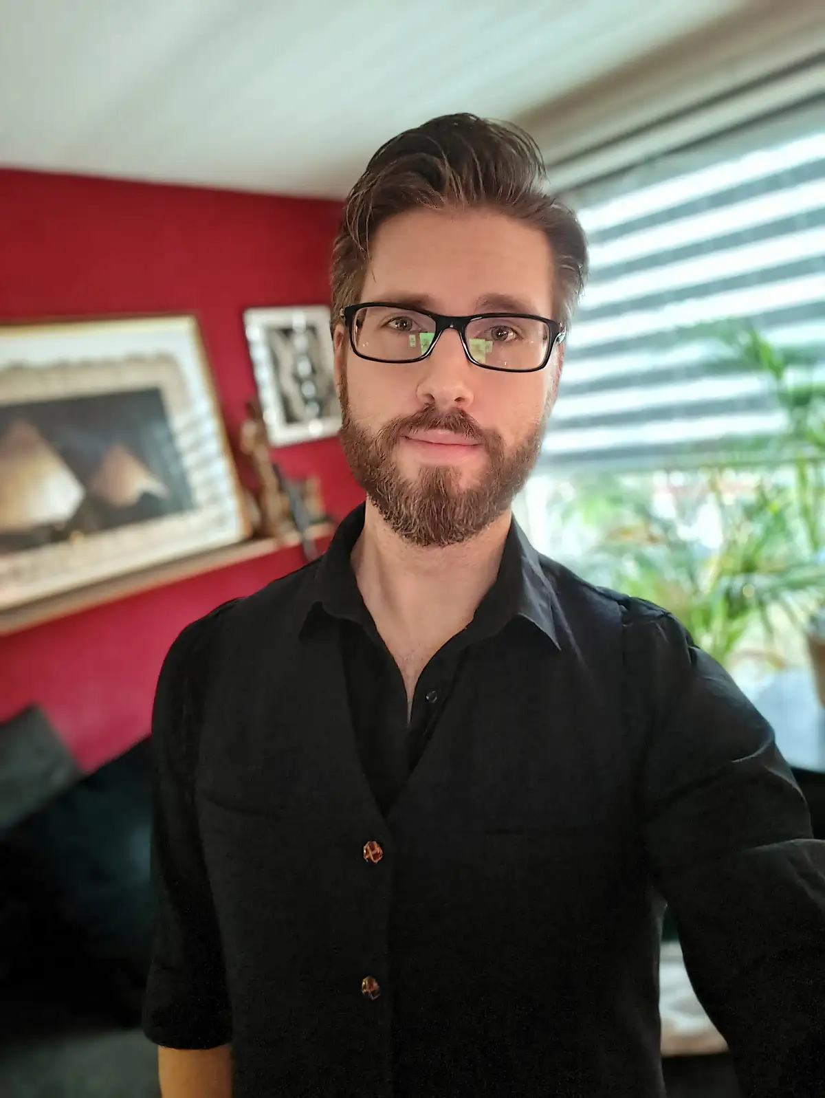 A man with a beard and glasses, wearing a black button-up shirt, stares directly at the camera with a neutral expression…