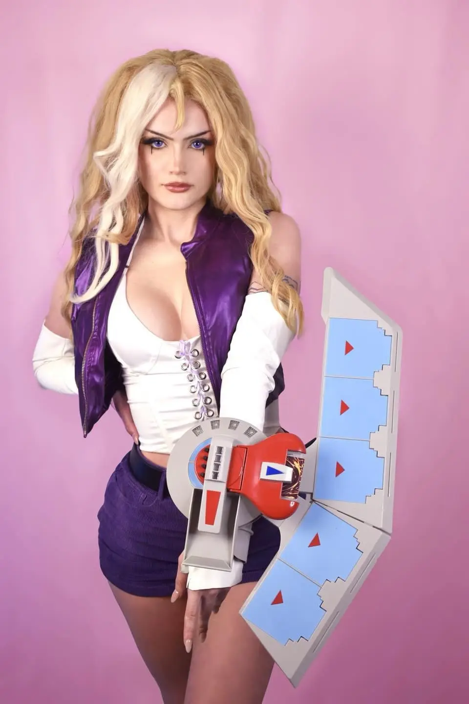 A person in cosplay as a blonde female character with purple eyes, wearing a purple vest over a white top and purple shorts…