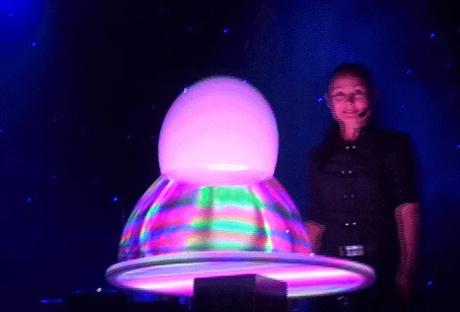 A magician performs a levitation trick with a glowing orb on a stage under blue lights. In the first panel, she smiles…