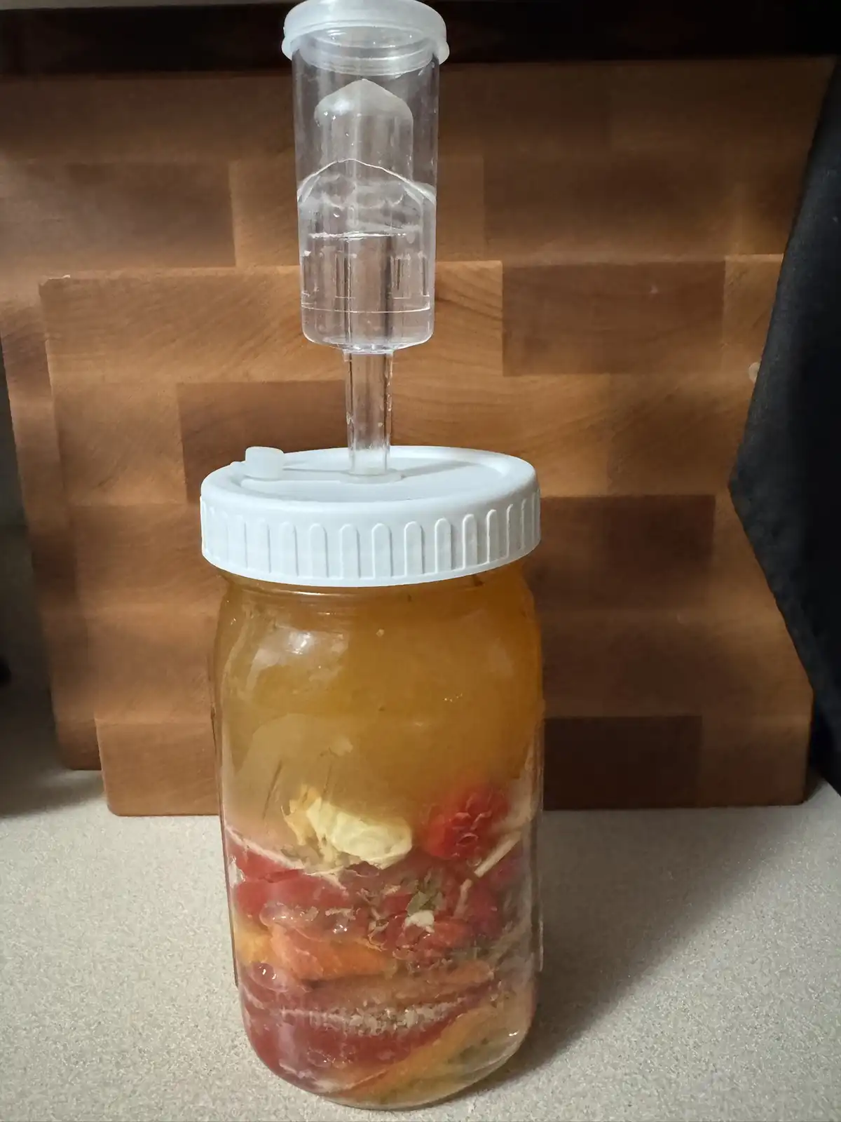 A glass mason jar filled with a cloudy amber liquid and visible chunks of fruit and herbs sits on a countertop, topped with…