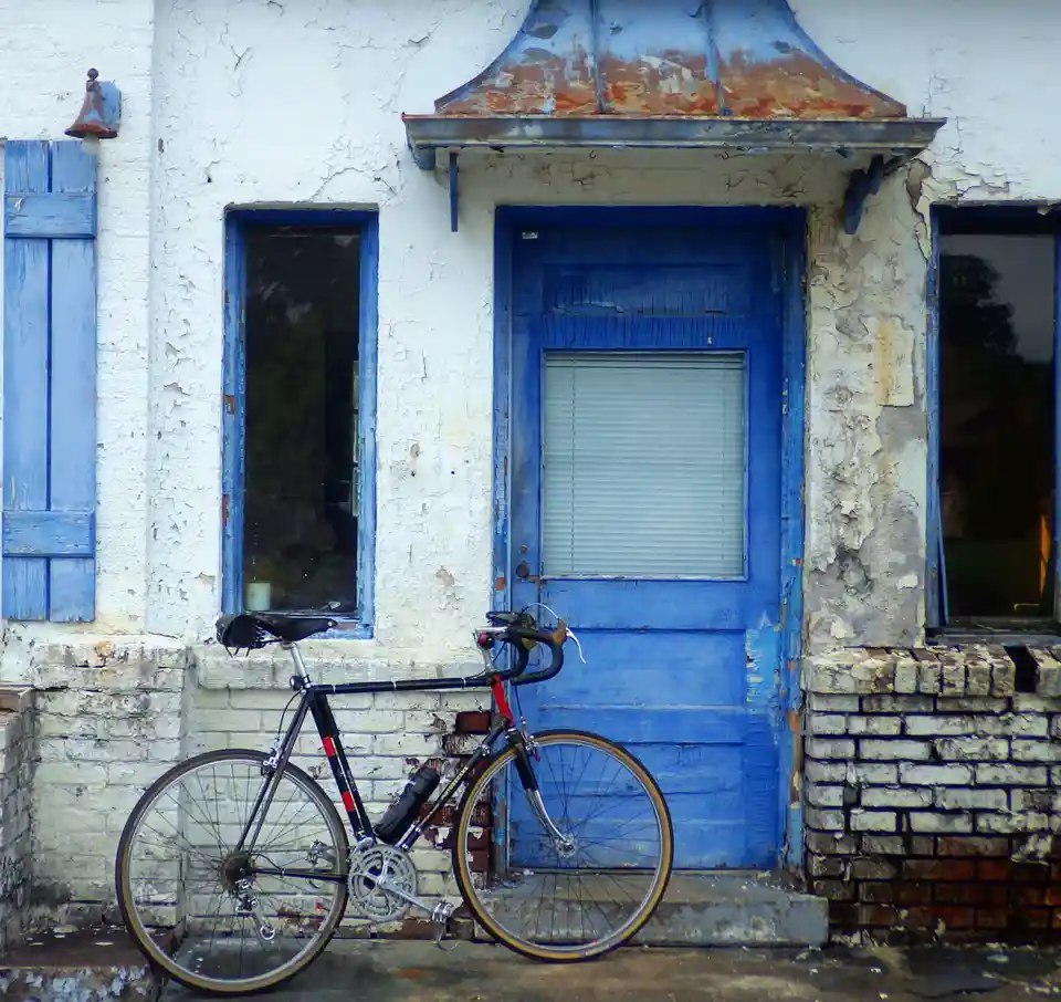 A black road bicycle with red accents is leaning against the steps of a dilapidated building with peeling white paint and a…