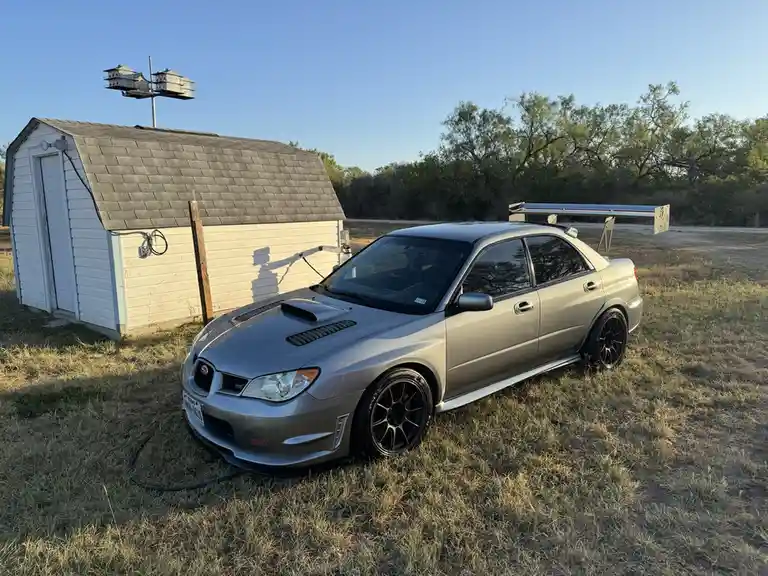 A silver Subaru WRX STI with a large rear wing and black wheels is parked on dry grass next to a small white shed. In the…